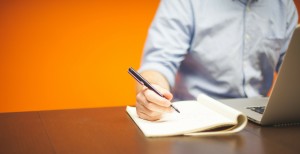 Man at desk with orange wall behind him copywriting on notepad
