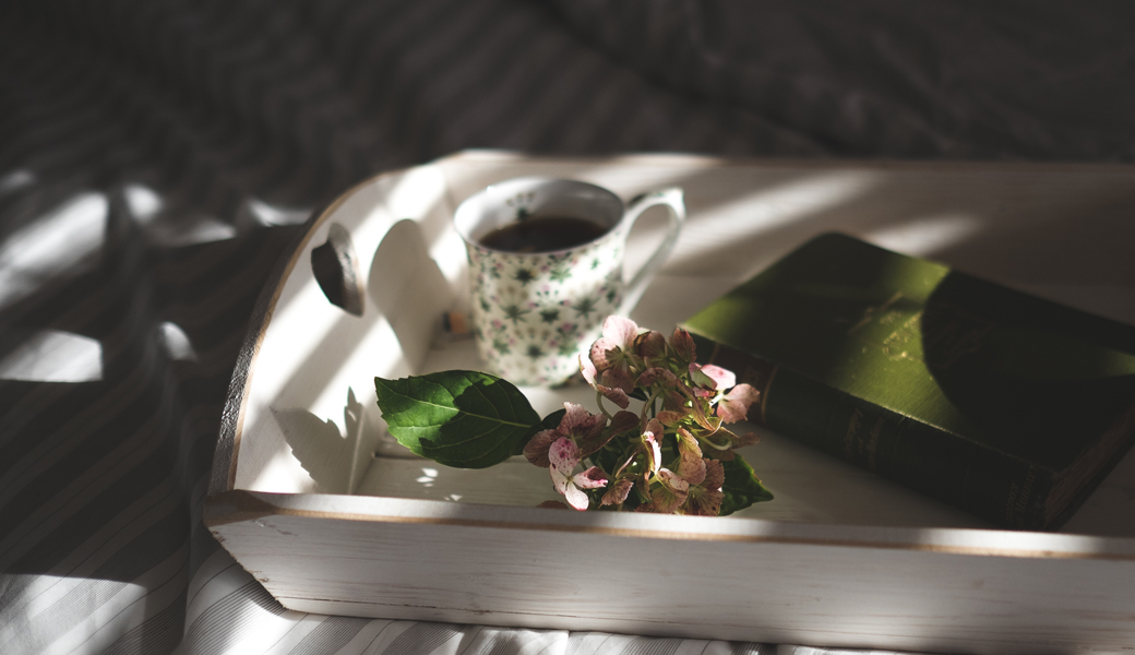 photo of a tray with a book coffee and some flowers resting on the sunlight in a bed